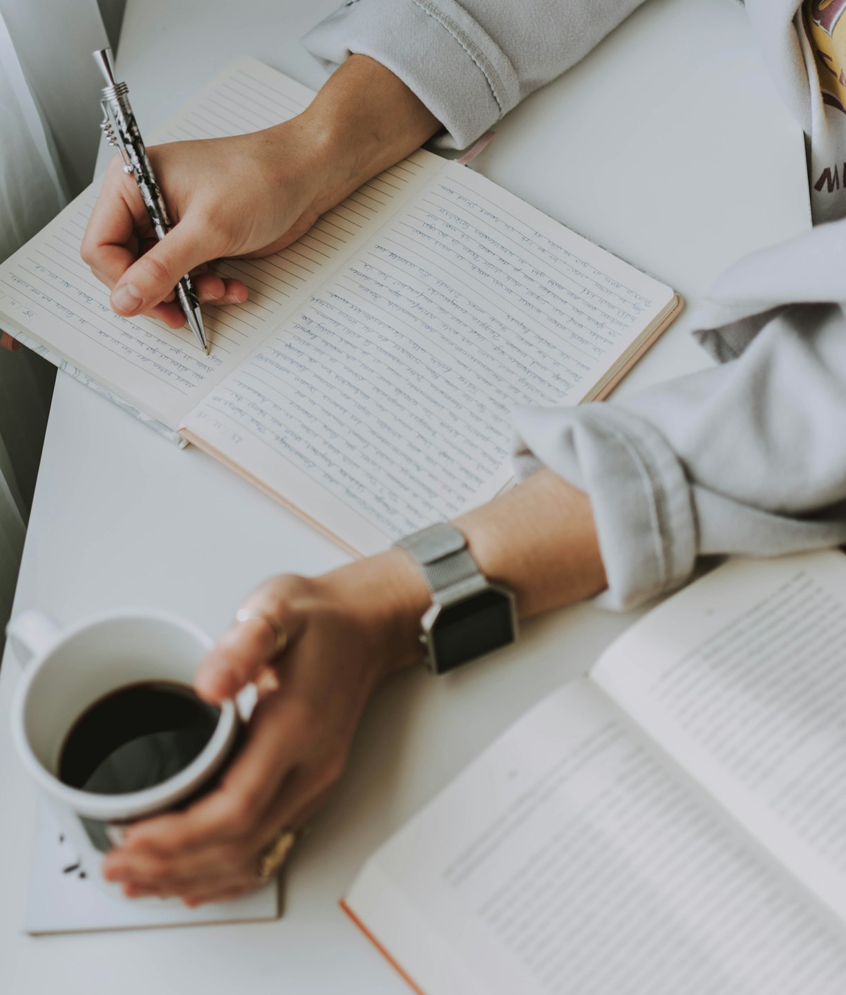 Hands writing in a notebook beside a cup of coffee.